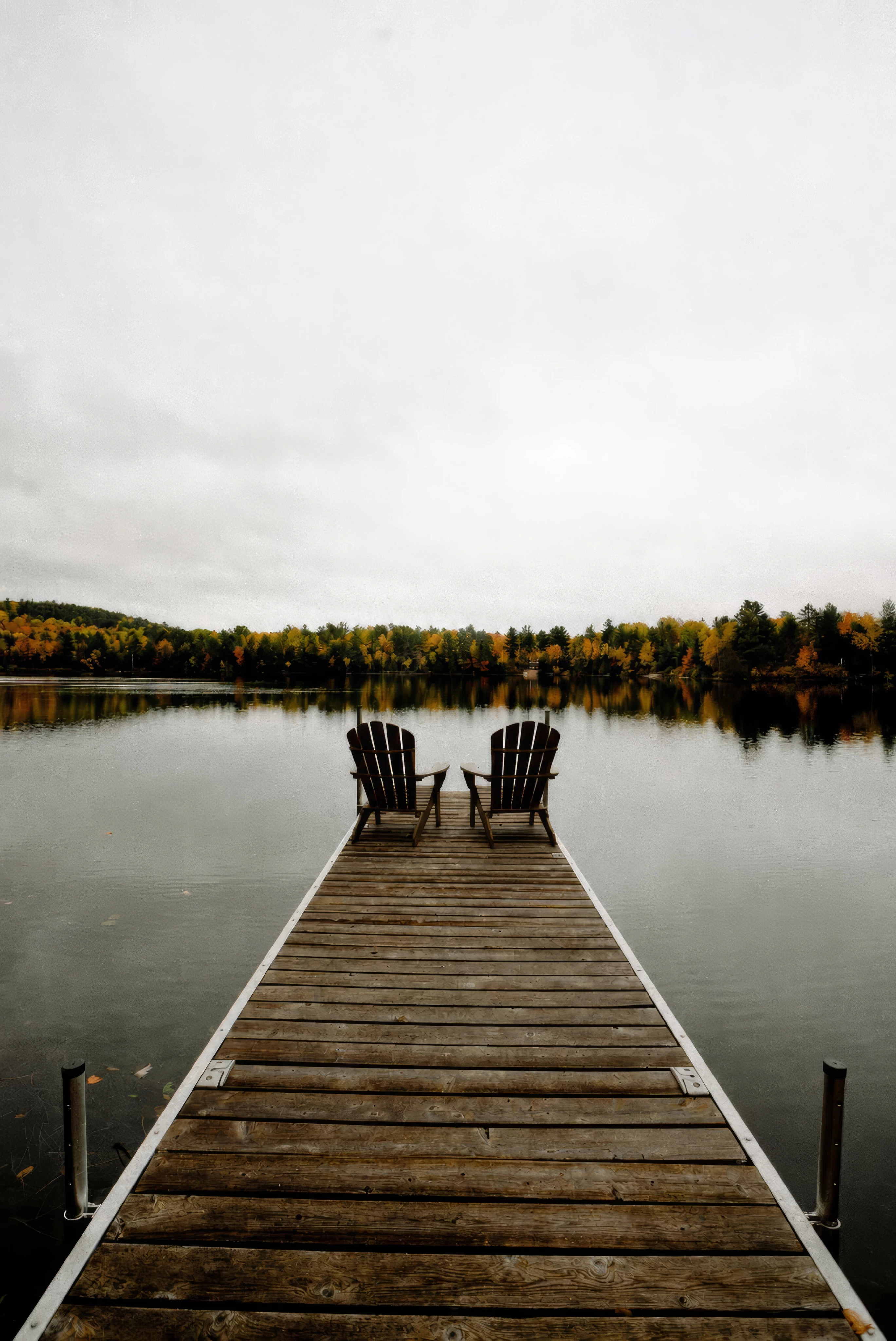Two empty Adirondack chairs at the end of a wooden dock overlooking a calm autumn lake.